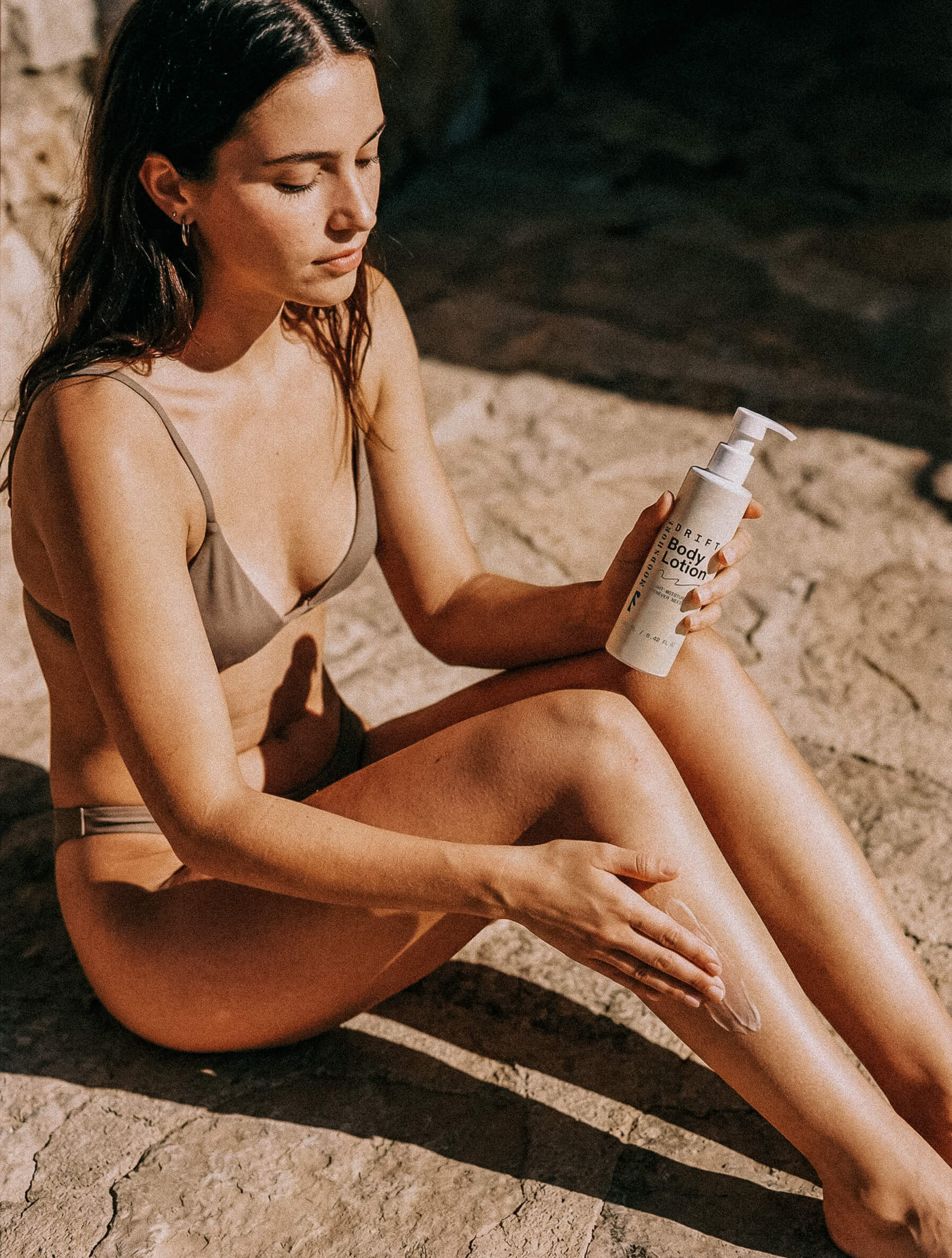 Woman applying Moodshore Drift Body Lotion to her leg on a rocky surface