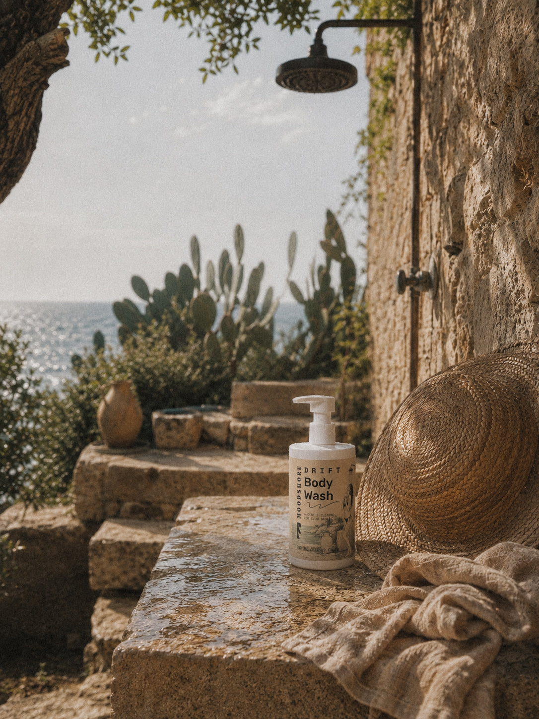 A bottle of Drift Body Wash sits on a stone ledge outdoors beside a woven straw hat and a beige towel, with a stone wall, shower fixture, cacti, and ocean in the background.